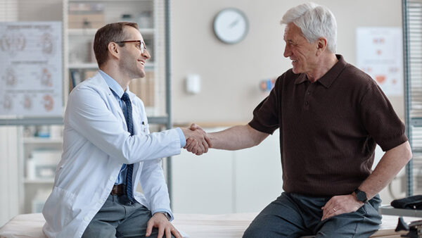 Middle aged Caucasian male doctor wearing glasses shaking hands with senior Caucasian man sitting on examination table in medical office during urology consultation